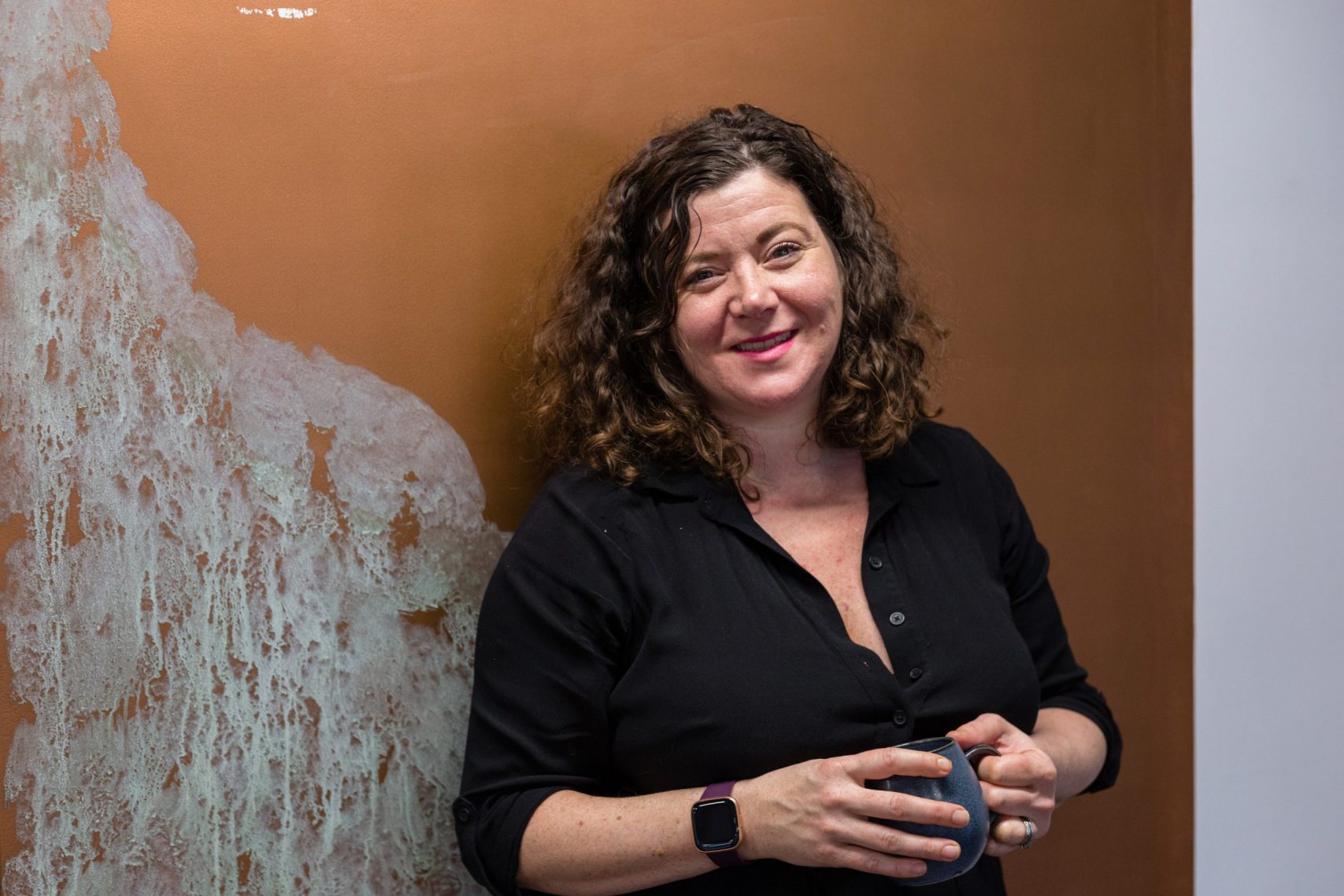 Melanie Johnson standing against a black and gold wall holding a mug of tea
